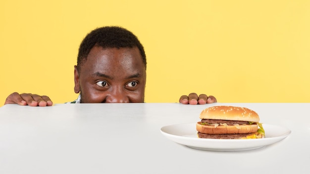 Hombre negro hambriento mirando Burger en mesa fondo amarillo