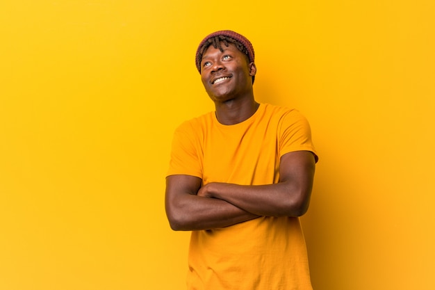 Hombre negro joven que lleva rastas sobre el fondo amarillo que sonríe confiado con los brazos cruzados.