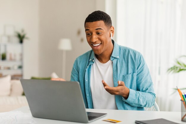 Hombre negro milenario sonriente en jeans con auriculares inalámbricos tiene videollamada en el interior de la oficina de la habitación