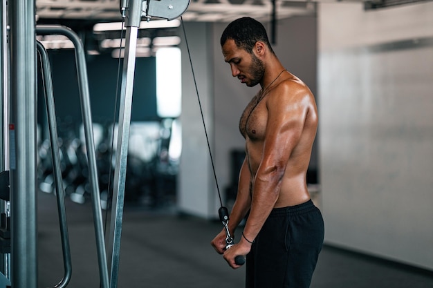 Hombre negro musculoso sin camisa entrenando con máquina cruzada en el gimnasio