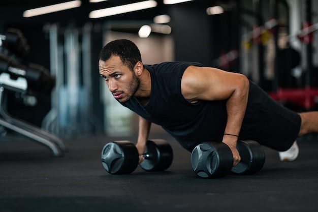 Hombre negro musculoso haciendo ejercicio de flexiones con mancuernas en el gimnasio