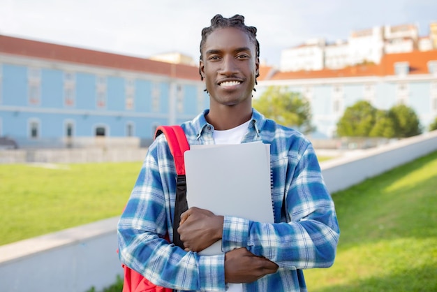 Hombre negro posa con libros y portátil en el parque del campus