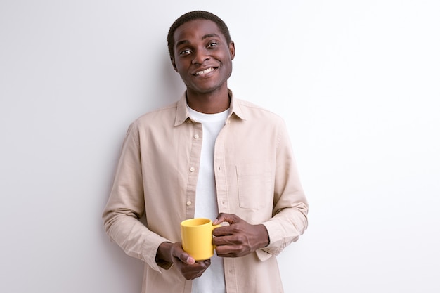 Hombre negro en ropa casual posando a la cámara sosteniendo la taza amarilla sonriendo. aislado en la pared blanca