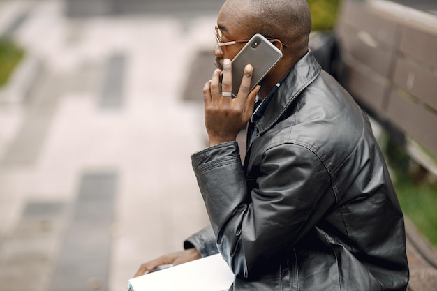 Hombre negro sentado en un banco en una ciudad. Hombre con gafas de sol y una chaqueta de cuero