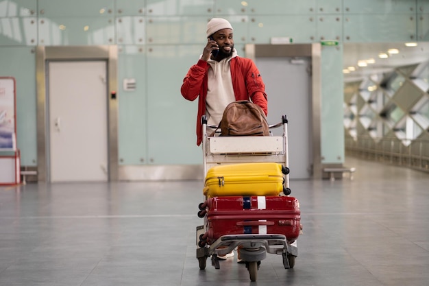 Hombre negro sonriente empujando el carrito de equipaje caminando después de la llegada al aeropuerto hablando por teléfono móvil