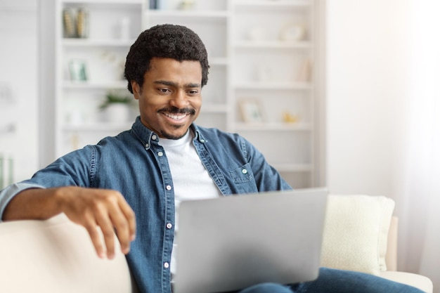 Hombre negro sonriente usando una computadora portátil en el sofá en una sala de estar brillante