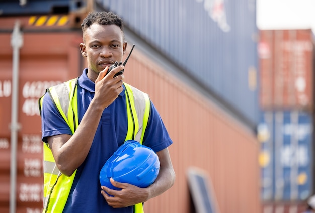 Hombre negro con walkie talkie en la terminal de contenedores, el trabajador industrial está controlando la carga