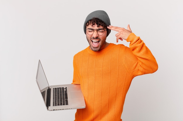 Hombre nerd con computadora que parece infeliz y estresado, gesto de suicidio haciendo un signo de pistola con la mano, apuntando a la cabeza