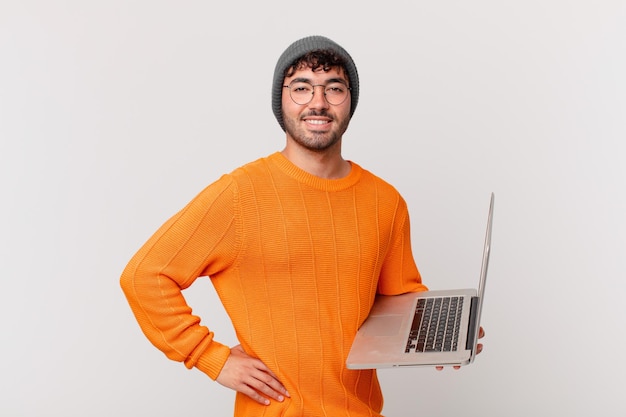 Hombre nerd con computadora sonriendo felizmente con una mano en la cadera y actitud confiada, positiva, orgullosa y amistosa