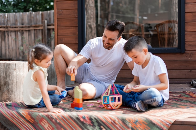 El hombre niñera juega con dos niños, sentado en el porche cerca de una casa de madera de campo, pasa el tiempo libre ayudando a los niños a recolectar cubos de juguete