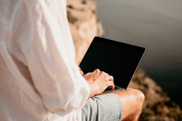 Hombre nómada digital en el sombrero un empresario con una computadora portátil se sienta en las rocas junto al mar durante la puesta de sol