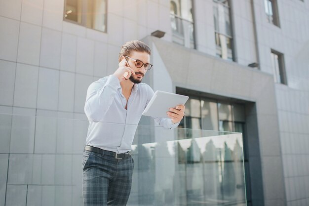 Hombre ocupado y guapo está parado y tiene tableta. Él mira su pantalla y habla por teléfono al mismo tiempo. Este chico es una persona multitarea.