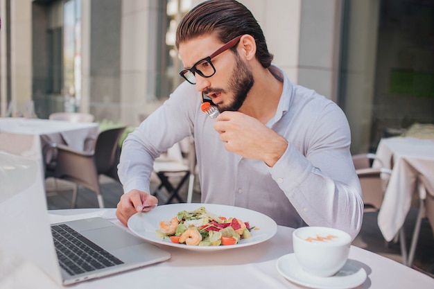 El hombre ocupado responde la llamada telefónica durante el almuerzo. Habla y sostiene el plato con ensalada al mismo tiempo. También hay una taza de café.