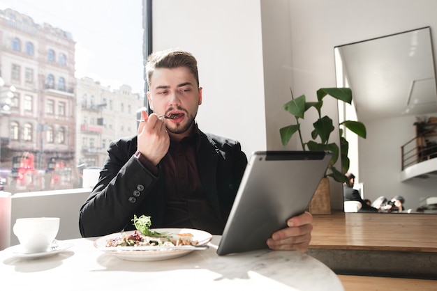 El hombre ocupado se sienta en un café, cena y mira videos en la tableta.