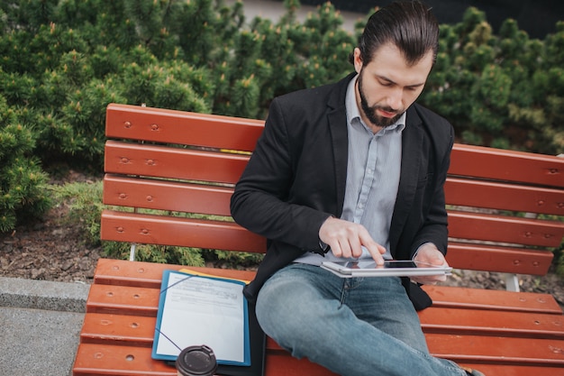 El hombre ocupado tiene prisa, no tiene tiempo, va a comer bocadillo al aire libre. Trabajador comiendo y trabajando con documentos en la tablet pc al mismo tiempo. Hombre de negocios realizando múltiples tareas.