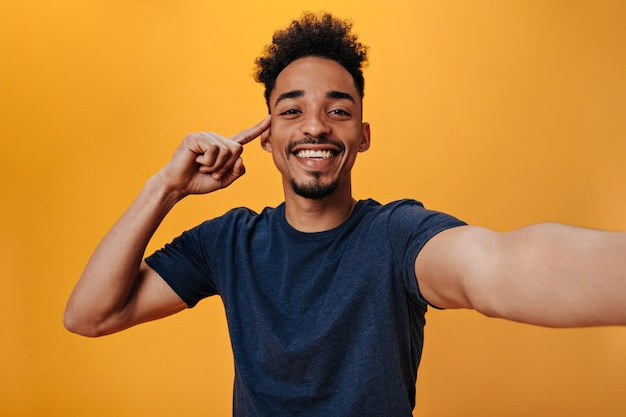 Hombre de ojos marrones en camiseta azul con sonrisa hace selfie en pared naranja