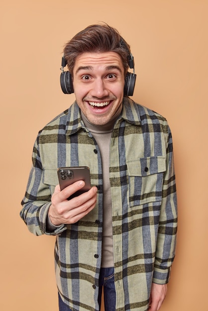 Hombre optimista positivo escucha pista de audio favorita en auriculares sostiene teléfono móvil reacciona en algo divertido vestido con camisa a cuadros casual aislado sobre fondo beige. Concepto de emociones