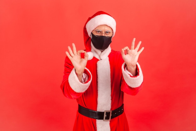 Hombre optimista positivo en traje de santa claus con máscara protectora que muestra un gesto correcto con los dedos animando la cuarentena Foto de estudio interior aislada sobre fondo rojo