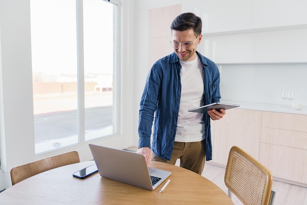 Un hombre optimista con ropa casual está de pie junto a una mesa con una computadora portátil sosteniendo una tableta en una habitación iluminada por el sol