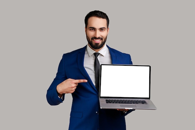 Hombre optimista señalando con el dedo a la computadora portátil con pantalla en blanco, mostrando una pantalla vacía, alardeando con publicidad en Internet, usando traje de estilo oficial. Disparo de estudio interior aislado sobre fondo gris.