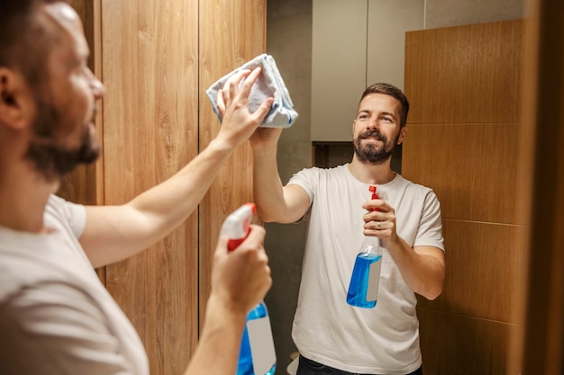 Un hombre ordenado está limpiando el espejo en un baño con detergente y tela.