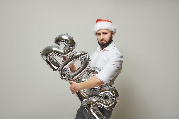 Un hombre orgulloso con un gorro de Papá Noel de terciopelo rojo sostiene globos plateados con la forma de 2022. Un hombre feliz con barba en una fiesta de año nuevo.