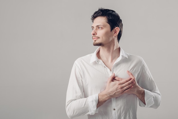 Hombre orgulloso joven hermoso en una camisa blanca que lleva a cabo la mano en el pecho en fondo gris.