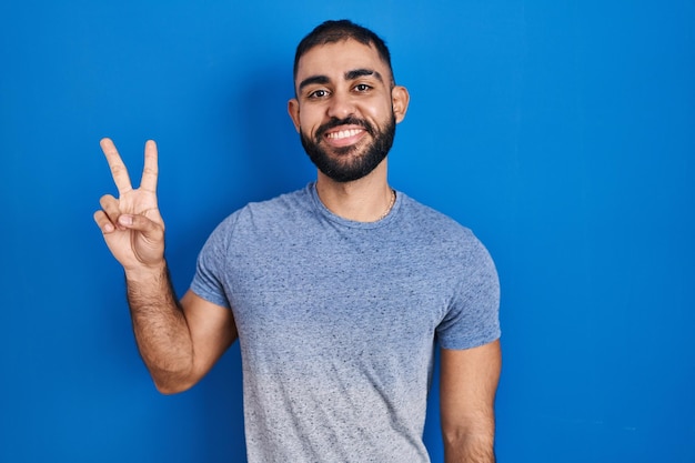 Hombre de Oriente Medio con barba sobre fondo azul sonriendo con cara feliz guiñando un ojo a la cámara haciendo el signo de la victoria con los dedos. número dos.