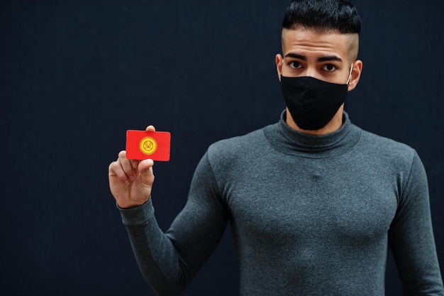 Hombre de Oriente Medio con cuello de tortuga gris y máscara de protección facial negra muestra antecedentes aislados de la bandera de Kirguistán