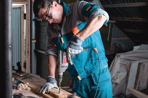 Hombre en overoles y gafas en el taller en un taladro, taladrar una tabla de madera