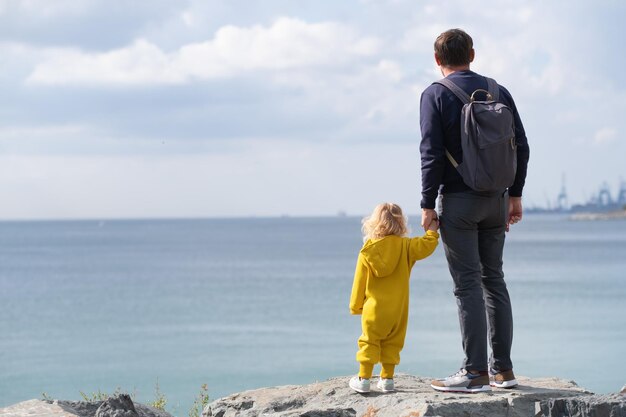 Hombre padre irreconocible sosteniendo a la niña pequeña mano mirando la hermosa vista al mar desde atrás feliz