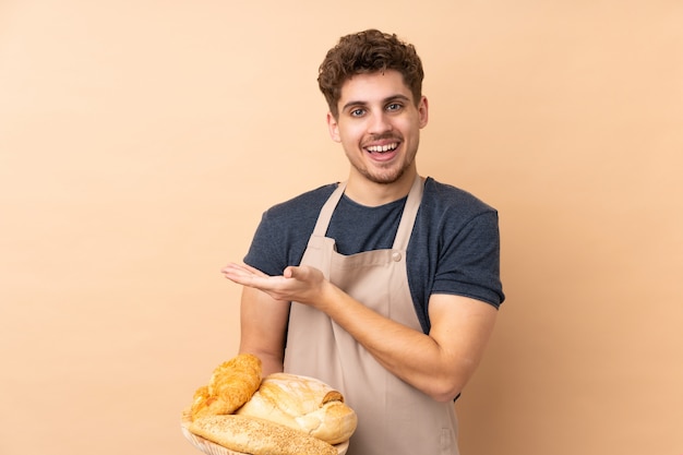 Hombre panadero sosteniendo una mesa con varios panes aislados en la pared beige extendiendo las manos a un lado para invitar a venir