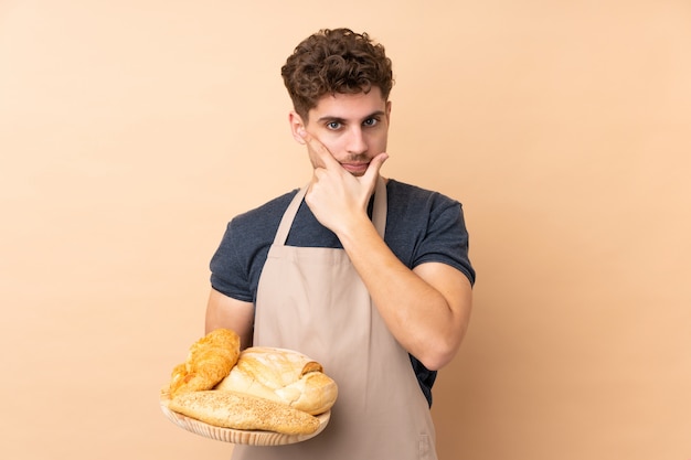 Hombre panadero sosteniendo una mesa con varios panes aislados en pared beige pensando en una idea