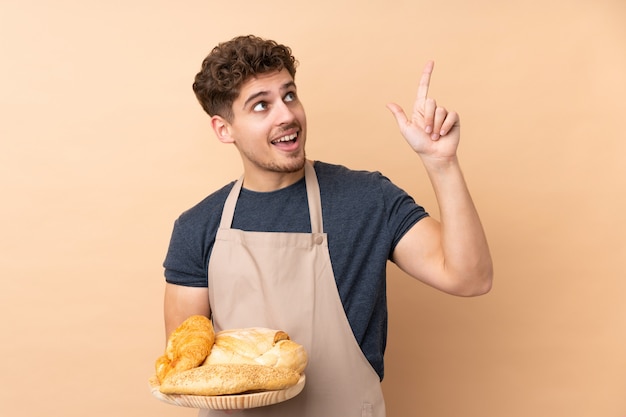 Hombre panadero sosteniendo una mesa con varios panes en la pared de color beige, señalando con el dedo índice una gran idea