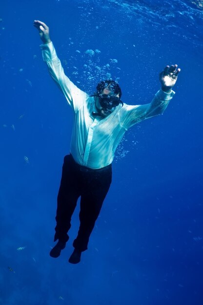 Foto hombre de pantalón negro, camisa blanca, hundiéndose lentamente en el abismo azul del mar.