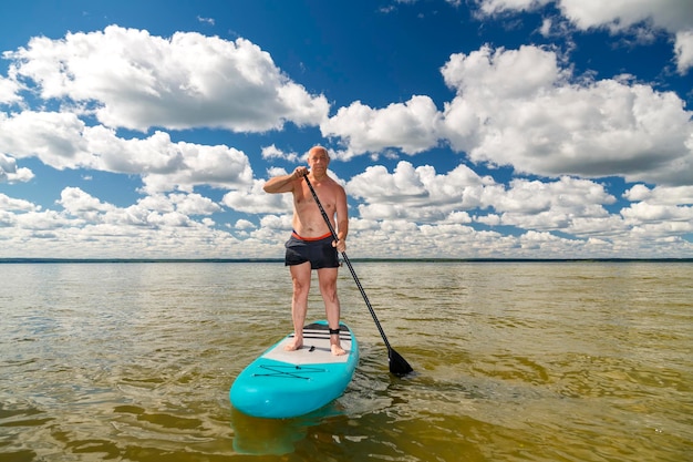 Foto un hombre en pantalones cortos deportivos con una paleta se para en una tabla sup en el lago contra el fondo de nubes blancas en un cielo azul claro