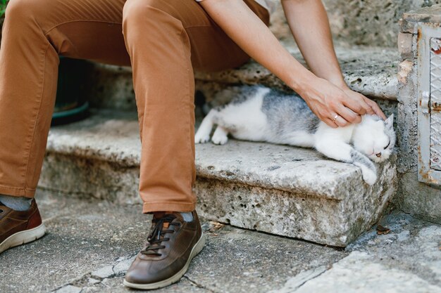 Un hombre con pantalones marrones se sienta en los escalones de piedra en el casco antiguo y acaricia un primer plano de gato gris acostado