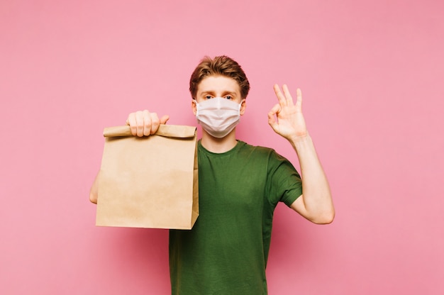 El hombre se para con un paquete de comida de la entrega en sus manos sobre una máscara médica en su rostro, mira a la cámara y muestra un gesto OK. Pandemia de coronavirus. Cuarentena. COVID-19.