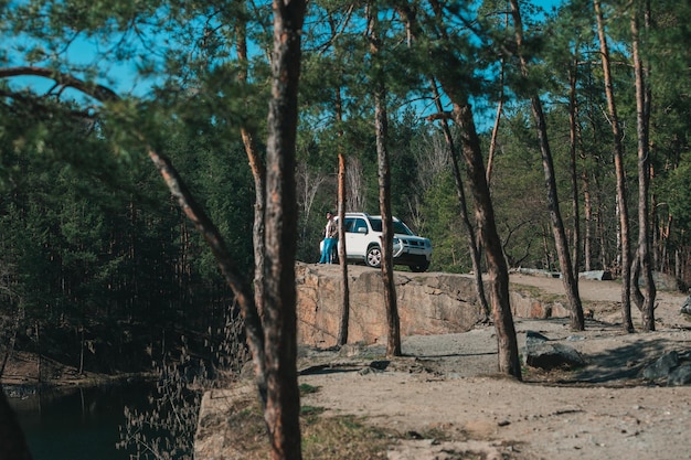 Hombre parado en un acantilado rocoso cerca de un auto todoterreno blanco con vista al lago a través de los árboles