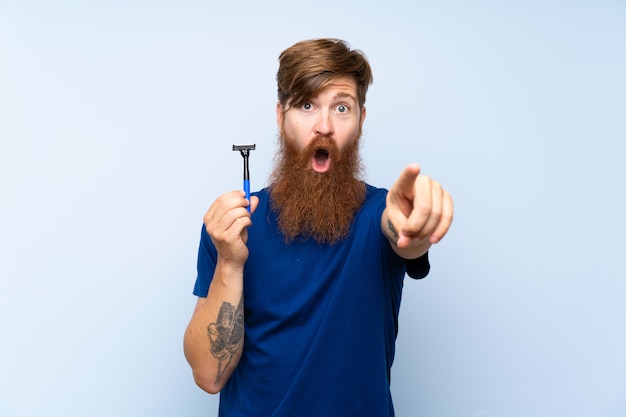 Hombre pelirrojo afeitarse la barba sobre la pared azul aislado sorprendido y apuntando al frente