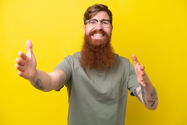 Hombre pelirrojo con barba aislado de fondo amarillo presentando e invitando a venir con la mano