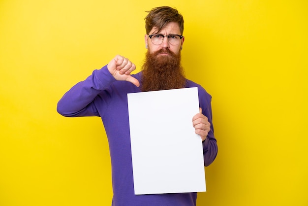 Hombre pelirrojo con barba aislado de fondo amarillo sosteniendo un cartel vacío y haciendo mala señal