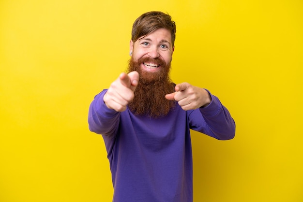 Hombre pelirrojo con barba aislado sobre fondo amarillo apuntando al frente con expresión feliz