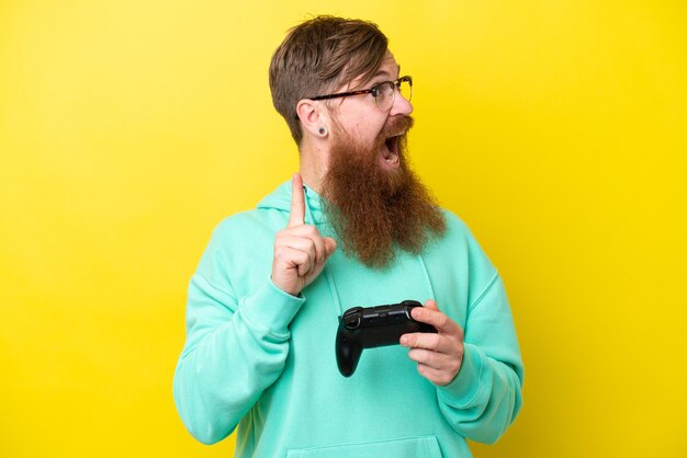 Hombre pelirrojo con barba jugando con un controlador de videojuegos aislado en un fondo amarillo con la intención de darse cuenta de la solución mientras levanta un dedo