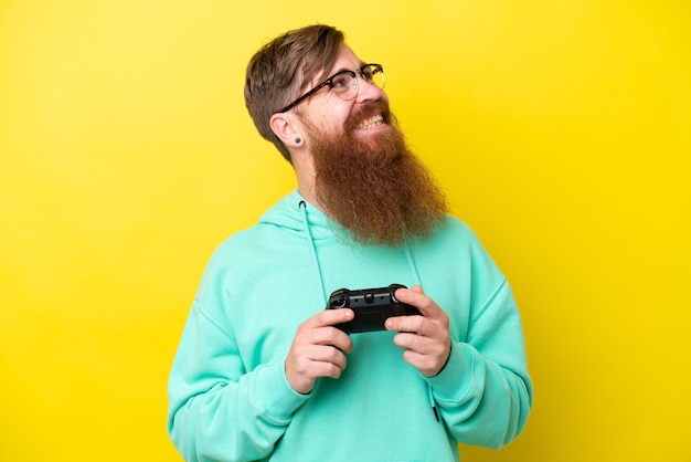 Hombre pelirrojo con barba jugando con un controlador de videojuegos aislado en un fondo amarillo mirando hacia arriba mientras sonríe