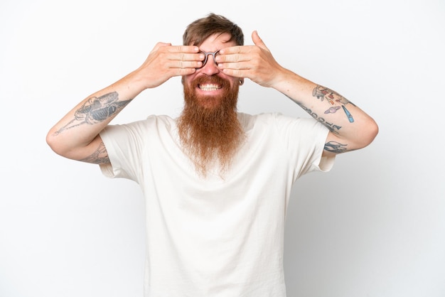 Hombre pelirrojo con barba larga aislado sobre fondo blanco cubriendo los ojos con las manos y sonriendo