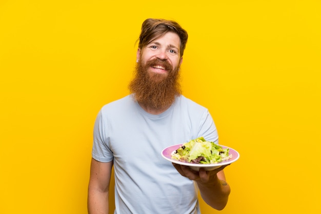 Hombre pelirrojo con barba larga y con ensalada sobre pared amarilla aislada sonriendo mucho