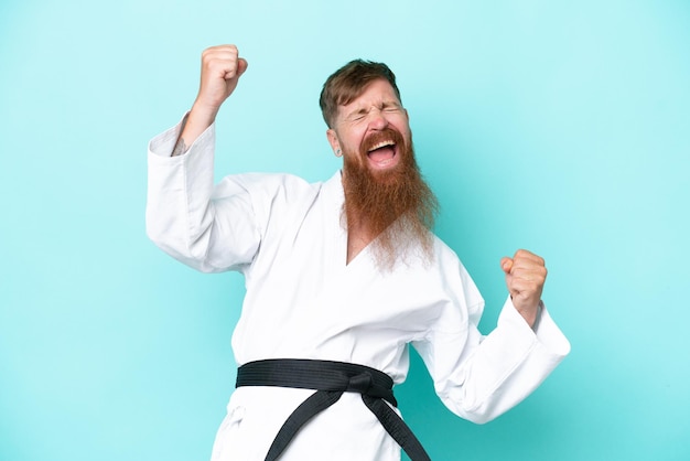 Hombre pelirrojo con barba larga haciendo karate aislado de fondo azul celebrando una victoria