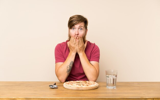 Foto hombre pelirrojo con barba larga en una mesa y con una pizza con expresión facial sorpresa