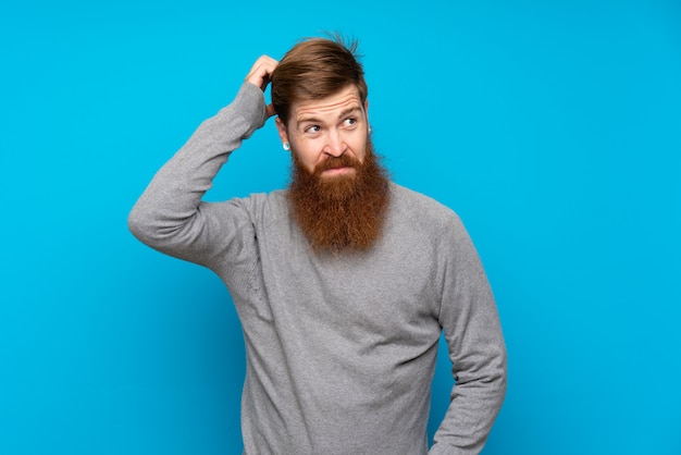 Hombre pelirrojo con barba larga sobre azul aislado que tiene dudas mientras se rasca la cabeza